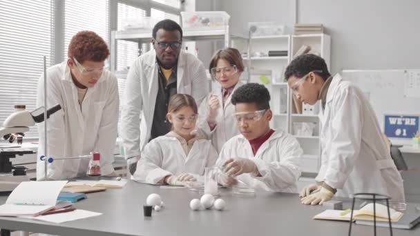 Lento grupo de estudiantes multiétnicos y su profesor afroamericano en batas de laboratorio experimentando con hielo seco consiguiendo mucho humo en frasco como reacción, divirtiéndose juntos en la clase de Ciencias - Metraje, vídeo