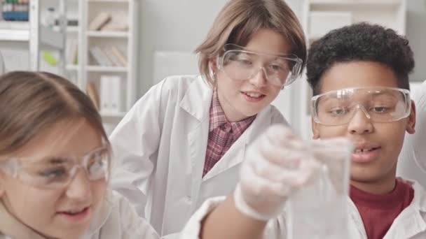 Cintura para cima tiro de escolares multiétnicos em jalecos e óculos de proteção conduzindo experimentos químicos na aula de Química. Biracial menino segurando frasco com água borbulhando nele - Filmagem, Vídeo