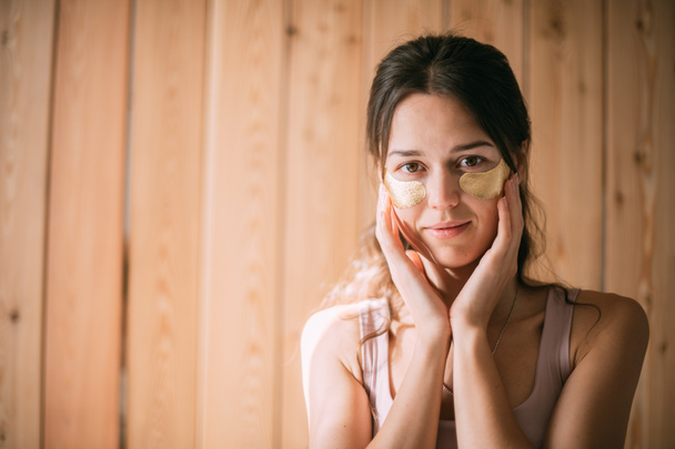 Portret van een jonge vrouw met gouden vlekken onder haar ogen tegen de achtergrond van een houten muur thuis. Mooie schattig brunette vrouw poseren op de camera met hyaluronische patches op een lichte achtergrond - Foto, afbeelding