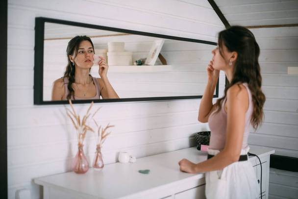 A young woman makes a facial massage with a gouache scraper at the mirror at home. A beautiful brunette makes facial skin care procedures with a gua sha stone near a large mirror in a bright room. - Zdjęcie, obraz