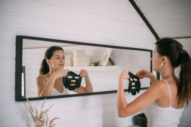 Portrait d'une jeune femme avec un masque noir au miroir à la maison. Une belle femme brune effectue des soins du visage avec un masque nettoyant et hydratant près du miroir dans une pièce lumineuse - Photo, image