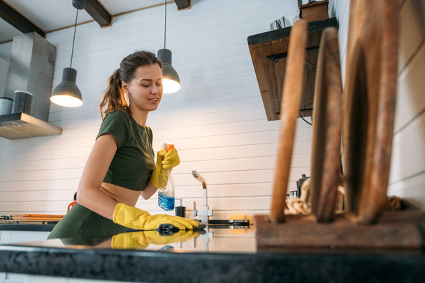 Weekend homework. Young pretty woman is cleaning the kitchen at home. A beautiful girl in home clothes and rubber gloves with a detergent in her hand washes the countertop in a large cozy kitchen - Foto, Imagen