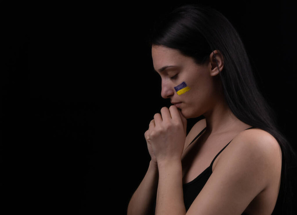 Portrait of serious ukrainian woman with blue and yellow ukrainian flag on her cheek. The symbol of the inviolability of the Ukrainian people. Dark background. Space for text - Foto, Imagen