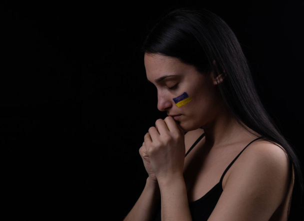Portrait of ukrainian woman with blue and yellow ukrainian flag on her cheek praying for Ukraine. The symbol of the inviolability of the Ukrainian people. Dark background. Space for text - Фото, изображение