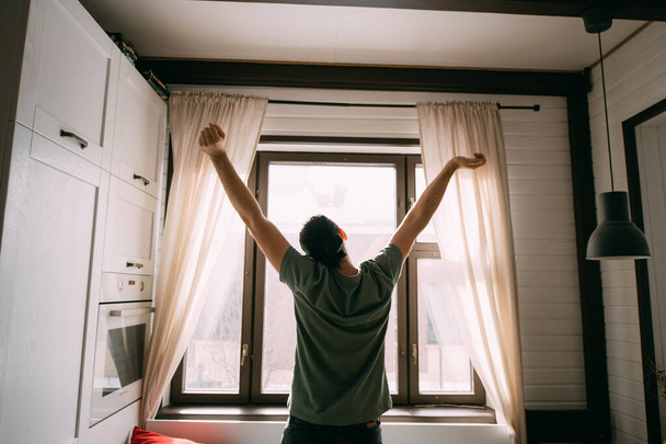 A fiatal elégedett férfi felhúzza a kezét a nappali ablakánál. Jóképű boldog srác nyújtózkodik reggel a nagy ablaknál egy napsütéses napon. - Fotó, kép