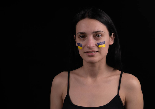 Portrait of young girl with blue and yellow ukrainian flag on her cheek. Dark background - Zdjęcie, obraz
