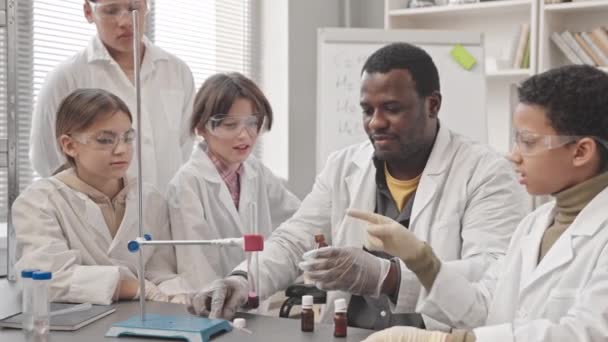 Střední záběr afroamerického muže Učitel chemie ukazuje laboratorní experiment žákům ve třídě, dává hnědý prášek do zkumavky s kapalinou a má kouř jako reakci - Záběry, video