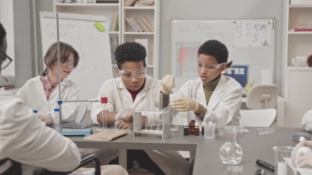 Média lentidão de alunas afro-americanas em óculos de proteção e jaleco fazendo experimentos químicos com tubos de laboratório sentados na mesa da aula de Ciências enquanto professores e colegas assistem - Filmagem, Vídeo