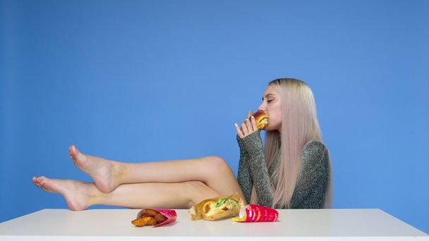Uma garota bonita senta-se em uma cadeira com as pernas na mesa e come um hambúrguer em um fundo azul. Dieta. O conceito de comida saudável e insalubre. fast food - Foto, Imagem
