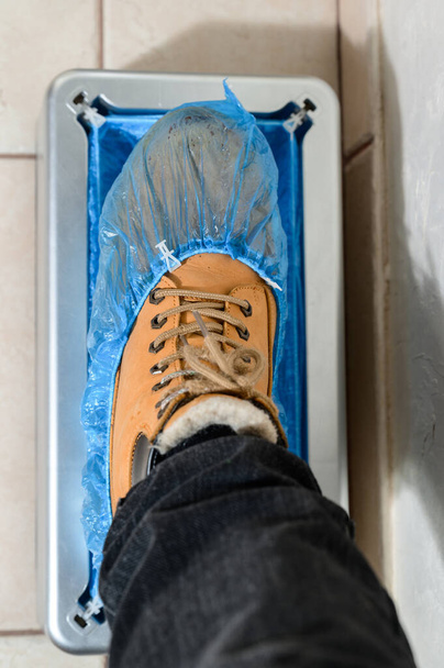 病院で使い捨ての靴カバーのための組織者である靴カバーとクリンプソッパー、男は部屋の清潔のための靴のブーツカバー、ブーツカバーを取り付けるための機械を置きます. - 写真・画像