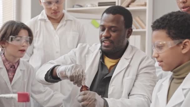 Colpo palmare di insegnante di scienza afroamericana che mostra esperimento di laboratorio agli scolari in camici e occhiali protettivi guardando tubo di vetro con fumo - Filmati, video