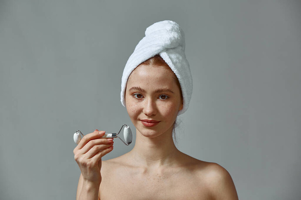 Young girl hold massager jade roller for facial massaging. Female with bare shoulders, towel on head show face massage - Foto, afbeelding