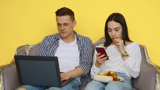 El chico y la chica comen comida rápida, el chico está sentado en un portátil, y la chica está mirando el teléfono en un fondo amarillo. El concepto de comida saludable y poco saludable. Comida rápida - Foto, Imagen
