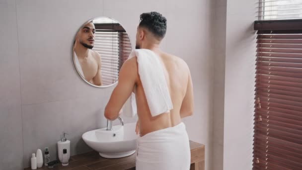 Desnudo árabe árabe indio barbudo hombre lavando la cara mirando en espejo limpiando la cara húmeda con toalla blanca chico mañana ducha cuidado de la piel rutina preparándose en baño belleza masculina cuidado de la piel lavado facial - Metraje, vídeo