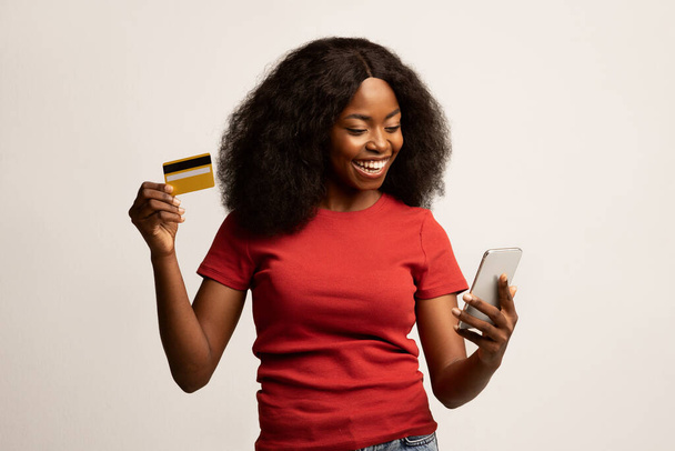 Compras en línea. Joven alegre hembra negra usando Smartphone y tarjeta de crédito - Foto, Imagen