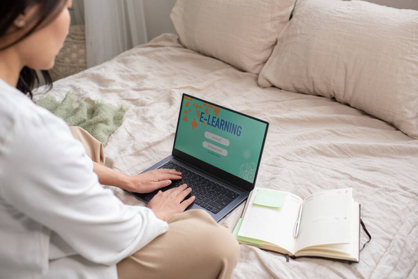 Молодая студентка в повседневной одежде сидит на кровати и вводит свое имя и пароль для входа на онлайн-курс обучения - Фото, изображение