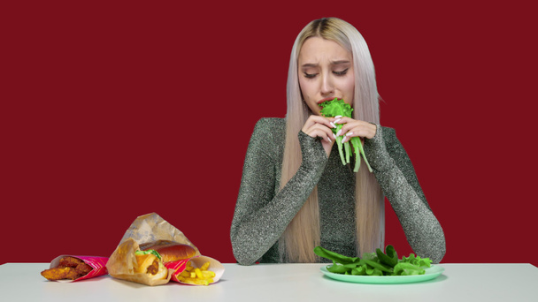 Egy aranyos lány zöldséget eszik, és szomorúan nézi a gyorskaját vörös alapon. Fogyókúra. Az egészséges és egészségtelen ételek fogalma. gyorsétterem - Fotó, kép
