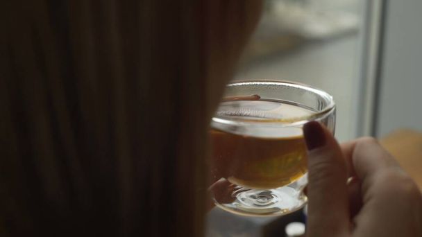 U okna sedí mladá dívka a drží skleněný hrnek s teplým čajem, který jí do šálku fouká studený vzduch. - Fotografie, Obrázek