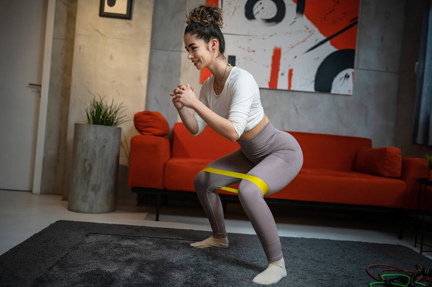 One woman beautiful female training at home in room using rubber resistance bands for legs squat and butt glutes sportswoman doing exercises alone health and fitness concept copy space full length - Foto, Imagen