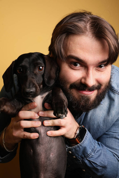 Photo of bearded young man with little dog posing over yellow background. - Zdjęcie, obraz