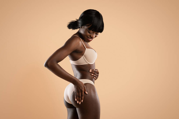 Улыбающаяся тощая африканская американка в нижнем белье, прикасающаяся к ягодицам - Фото, изображение