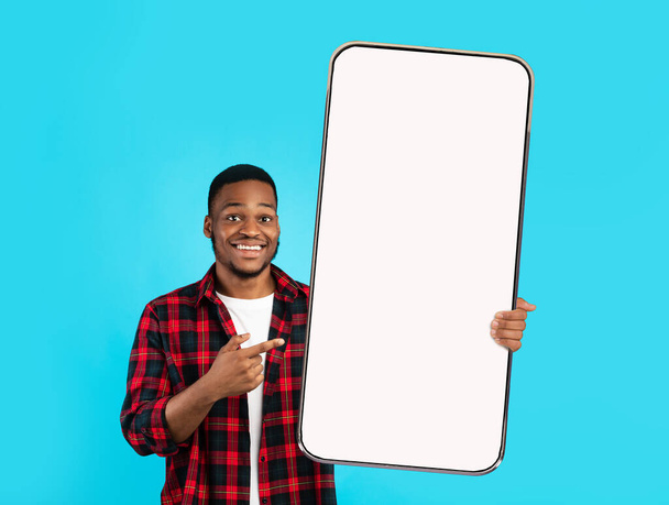 Έκπληκτος χαρούμενος νεαρός αφροαμερικάνος τύπος σε casual σημεία δάχτυλο σε τεράστιο smartphone με λευκή οθόνη - Φωτογραφία, εικόνα