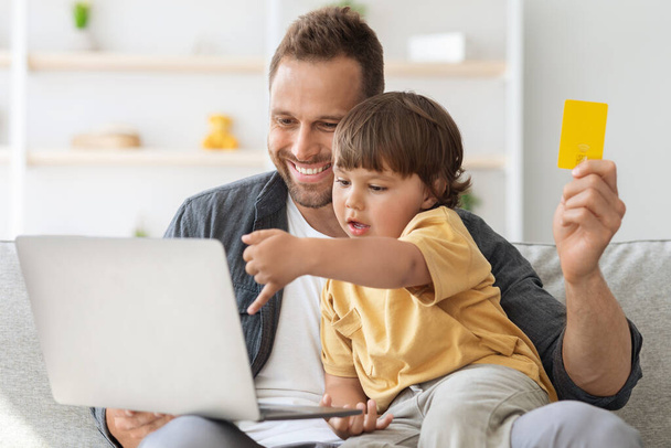 Інтернет-магазини. Щасливий молодий батько робить інтернет покупки зі своїм маленьким сином, хлопчик вибирає іграшки на ноутбуці
 - Фото, зображення