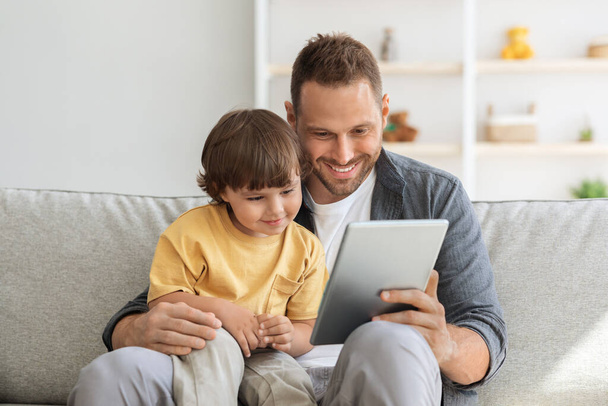Çocuk gelişimi içeriği. Genç baba, küçük sevimli oğlana dijital tablet gösteriyor. Çocuk internette eğitim programından hoşlanıyor. - Fotoğraf, Görsel