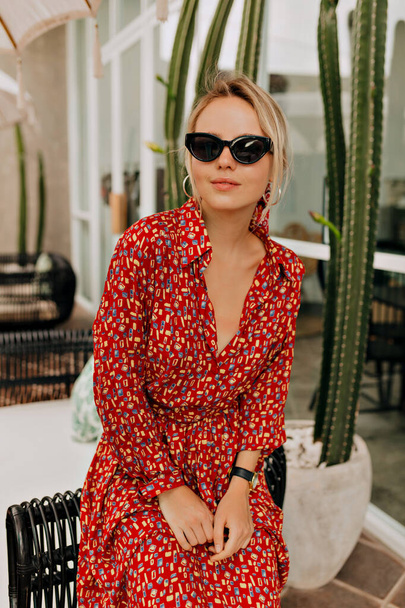 Sarışın, sevimli bir kadın, güneş gözlüğü takıyor ve parlak renkli bir yaz elbisesi giyiyor. Kafeterya yakınlarında egzotik bitkilerin arka planında dinlenirken kameraya mutlu bir gülümsemeyle poz veriyor. - Fotoğraf, Görsel