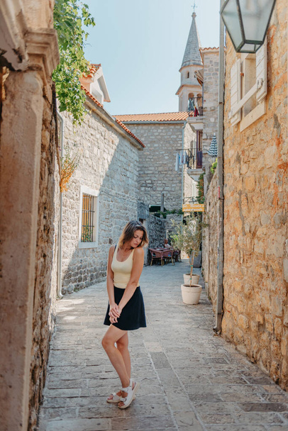 Girl Tourist Walking Through Ancient Narrow Street On A Beautiful Summer Day In MEDITERRANEAN MEDIEVAL CITY, OLD TOWN BUDVA, MONTENEGRO. Young Beautiful Cheerful Woman Walking On Old Street At - Φωτογραφία, εικόνα