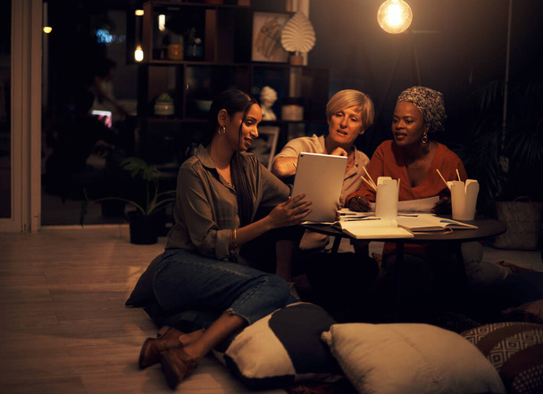 This app will help us complete the deadline. Shot of a group of businesswomen working together on a digital tablet in an office at night. - Foto, imagen
