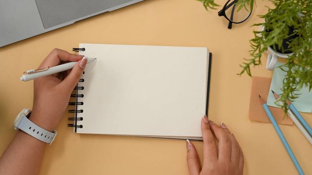 Vrouwelijke universiteitsstudent maakt aantekeningen op spiraalvormig notitieblok, schrijft een wenskaart op haar werkruimte. Lege spiraalvormige notitieblok model. bovenaanzicht - Foto, afbeelding
