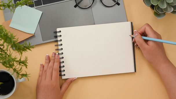 Female officer wiring her ideas on spiral notepad in her minimal office desk. Empty spiral notepad mockup. top view  - Фото, изображение
