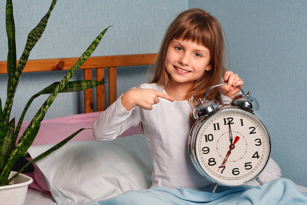 Огромный будильник в руках счастливой улыбающейся девочки в спальне. 7 часов утра.. - Фото, изображение