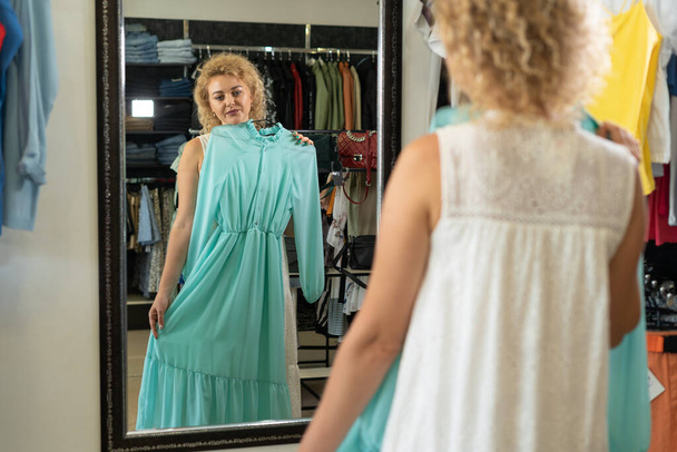 Женщина примеряет платье перед зеркалом в бутике, помещение для примерки в магазине одежды, отдел женской одежды торгового центра, девушка выбирает летнее хлопковое платье, концепция покупок - Фото, изображение