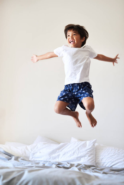Πετάω μόνος. Πλήρες πλάνο ενός αξιολάτρευτου μικρού αγοριού που παίζει και πηδάει σε ένα κρεβάτι στο σπίτι. - Φωτογραφία, εικόνα