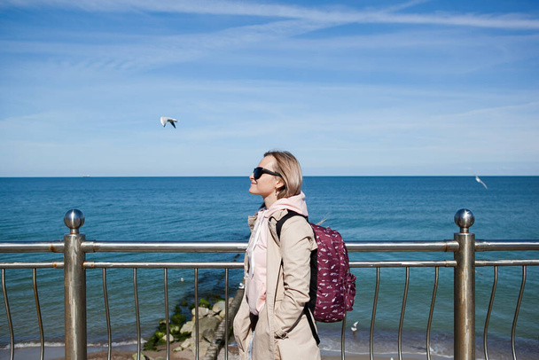 a blonde girl with a backpack on the background of a cold sea, seagulls in the sky. the concept of travel, trips, immigration - Foto, afbeelding