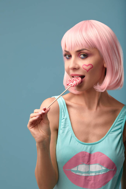 Großaufnahme eines koketten weiblichen Models auf blauem Hintergrund, das eine rosa Perücke trägt und verschiedene Arten von Bonbons in der Hand hält. Ziemlich glamouröse Frau rosa Haare Charme Süßigkeiten Lifestyle Genuss Zucker - Foto, Bild