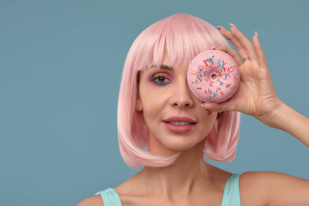 Pembe peruklu, elinde pembe bir donutla gözünün önünden ayırmayan, dilini gösteren şık bir kızın portresi. - Fotoğraf, Görsel