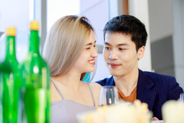 数百万人の若い恋人カップルアジアの夫で正式なスーツと美しい妻笑顔開催背の高いグラスワイン乾杯一緒に幸せな新年パーティーで装飾された飲料と食品テーブル. - 写真・画像