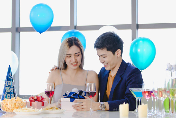Millennial joven amante pareja asiática marido en traje formal y hermosa esposa sonriendo sosteniendo vasos altos vino brindis juntos en feliz fiesta de año nuevo en la mesa de bebidas y alimentos decorados. - Foto, imagen
