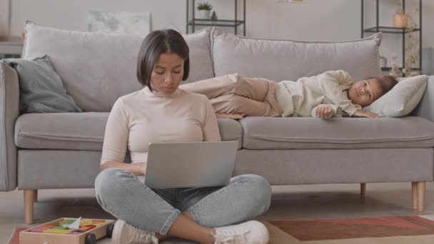Lassú fiatal Biracial nő dolgozik laptop ül szőnyegen a nappaliban, míg az unatkozó 8 éves lánya zavarja őt a munka akar játszani, együtt maradni otthon zárlat alatt - Felvétel, videó