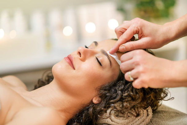 Beauty specialist verwijdert haar van wenkbrauwen met wax strips aan rustige klant met gesloten ogen liggend op de bank in beauty studio - Foto, afbeelding