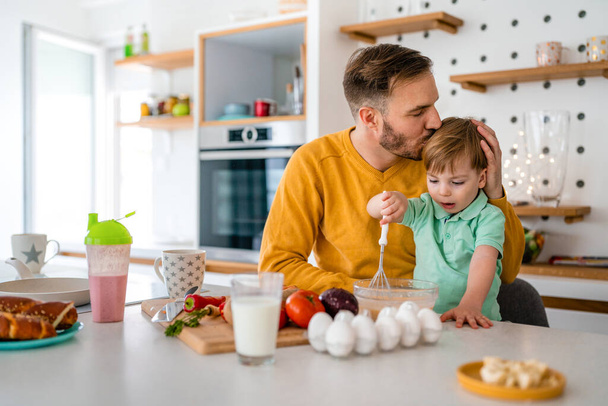 Young happy single father with a toddler boy having fun and preparing healthy food in kitchen at home. - Fotoğraf, Görsel