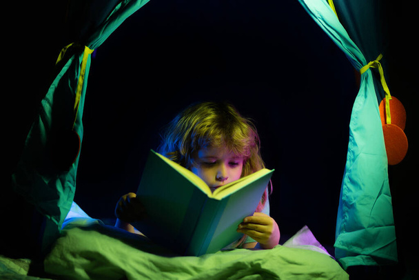 Kids in kids tent reading a book in the dark home. Child boy reading a book lying on the bed. Dreaming child read bedtime stories, fairystory or fairytale. - Photo, image