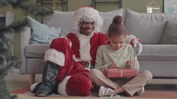 Slowmo di bella ragazza afroamericana di 8 anni che scarta la scatola regalo da Babbo Natale seduti insieme sul pavimento dal divano in soggiorno - Filmati, video