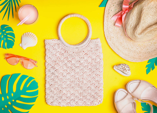 Summer female outfit. Flat lay.  Woven hand bag, straw hat, sunglasses, sandals with cocktail , seashells and tropical paper palm leaves on yellow background, top view. Summer holiday vacation. - Foto, Imagem