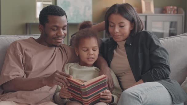 Keskipitkä hidas melko 8-vuotias afroamerikkalainen tyttö istuu sohvalla rakastavien vanhempiensa välillä lukemassa kirjaa yhdessä vapaa-ajalla kotona - Materiaali, video