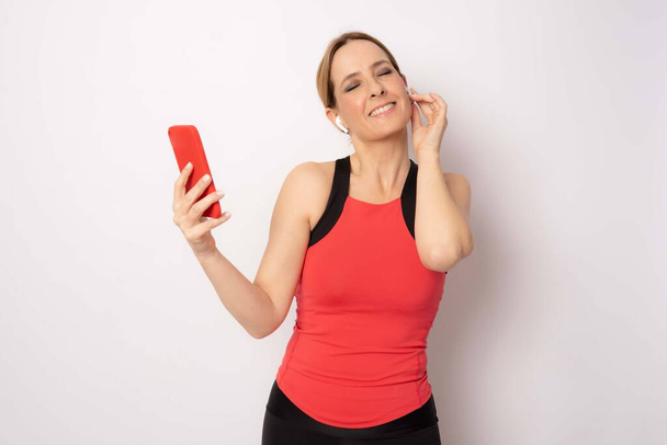 Mujer joven feliz en ropa deportiva sonriendo y sosteniendo el teléfono inteligente y escuchar música sobre fondo blanco. - Foto, imagen
