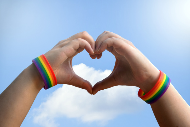 lgbt, same-sex love and homosexual relationships concept - close up of male hands with gay pride rainbow awareness wristbands showing heart gesture with bluesky background. - Photo, Image